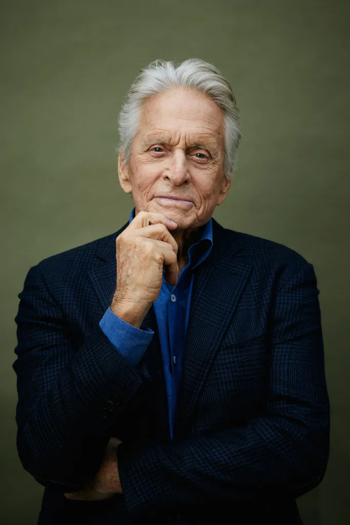 Portrait of Michael Douglas with his hand on his chin, wearing a blue shirt and dark blazer against a green background.