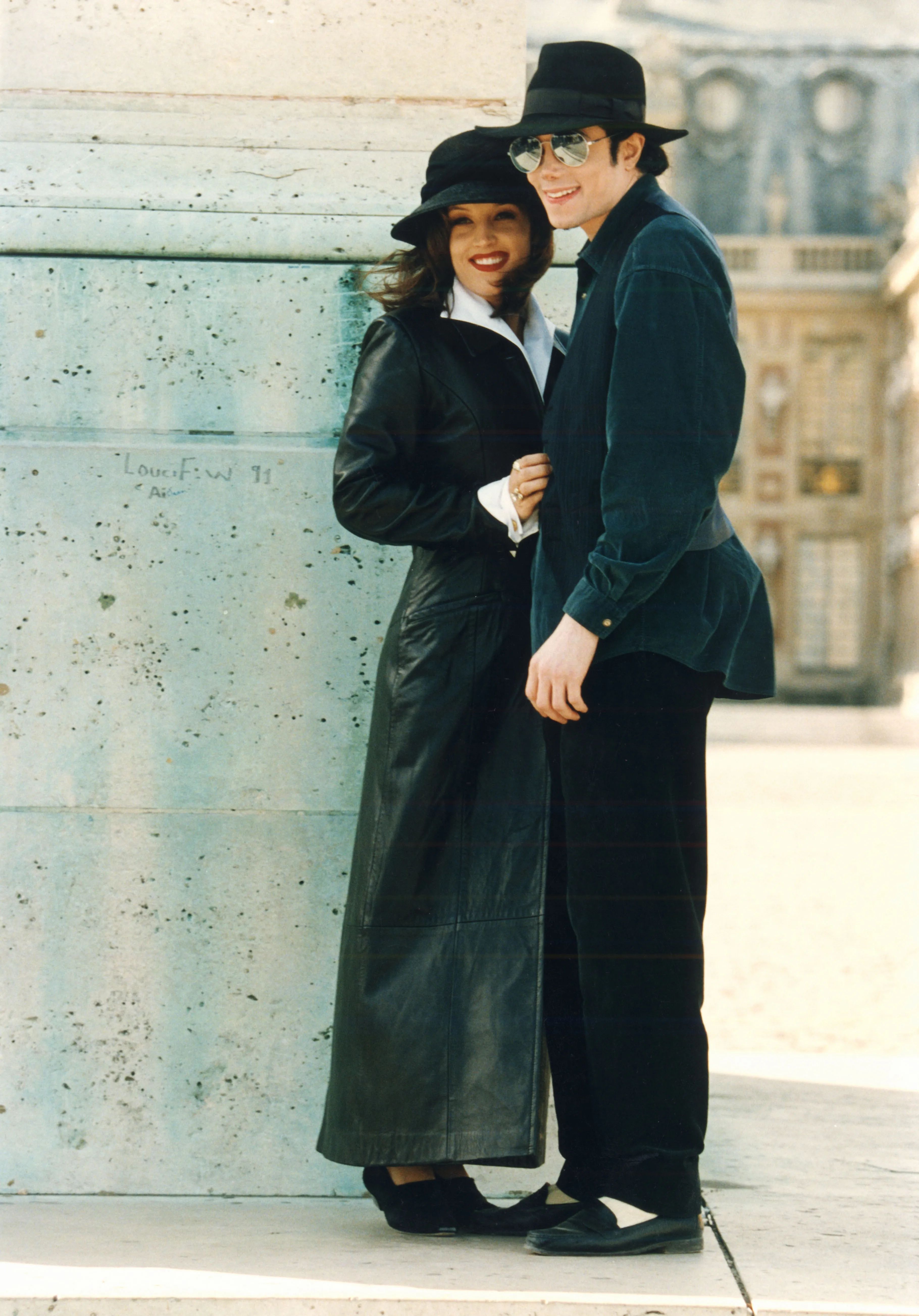 Michael Jackson and Lisa Marie Presley standing together outdoors near a large pillar, both wearing black hats and dark clothing.