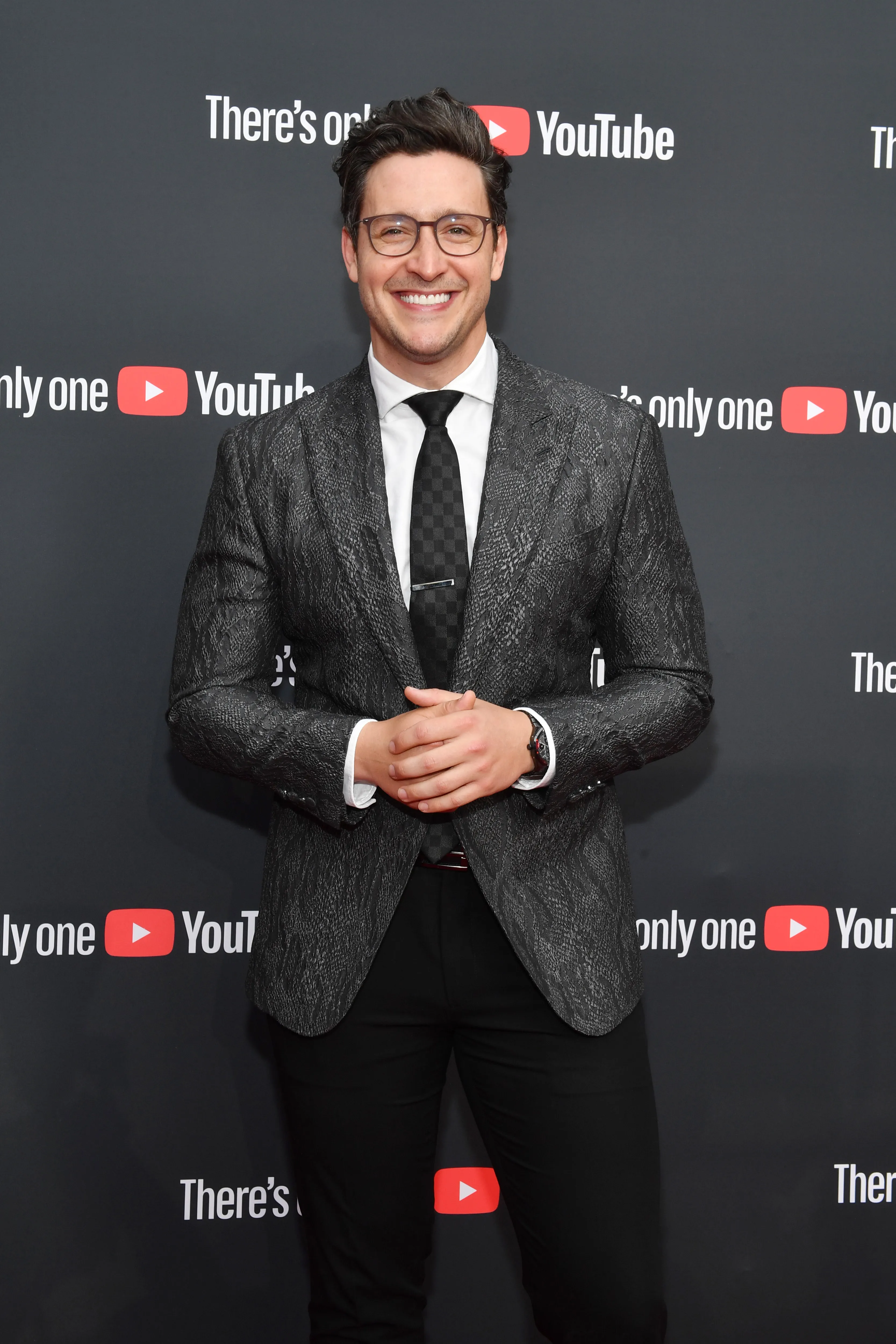 Dr. Mike Varshavski smiling in a patterned grey suit, white shirt, and black tie in front of a YouTube Brandcast backdrop.