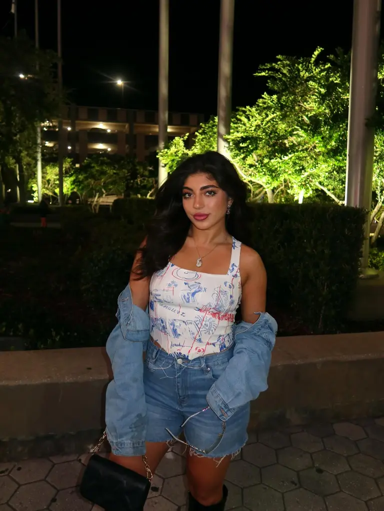 Milania Giudice posing outdoors at night in a patterned top, denim shorts, and an off-the-shoulder denim jacket.