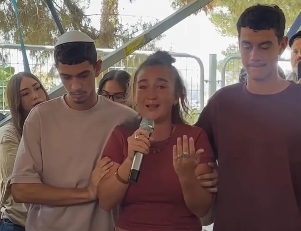 Miriam Ohayon speaks into a microphone while flanked by people at her brother's memorial.
