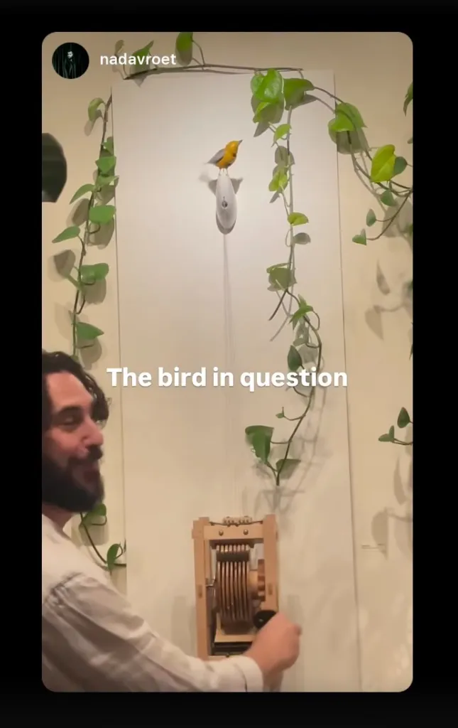 A man operating the crank of a mechanical sculpture featuring a yellow animated bird.