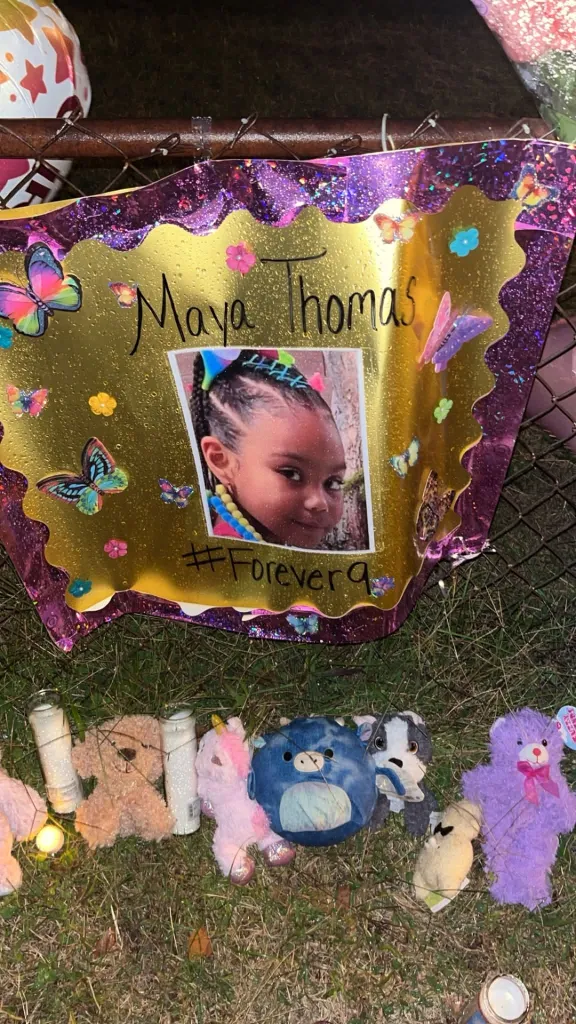 A memorial for Maya Thomas features a golden sign with her picture, surrounded by stuffed animals and lit candles.
