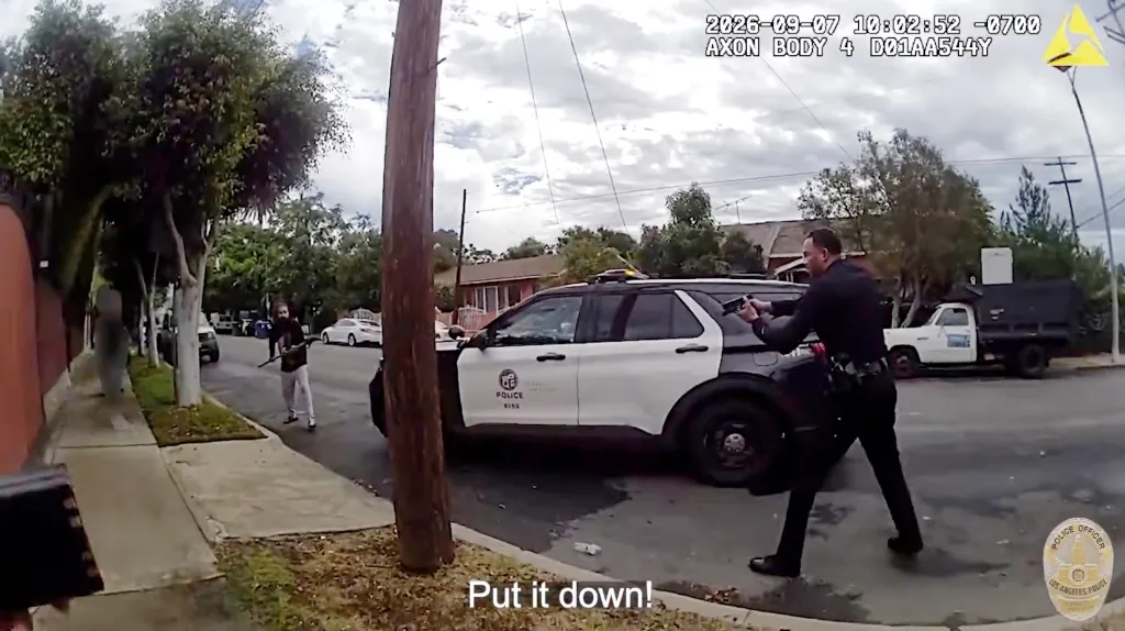 Bodycam footage of a police officer pointing a gun at a man holding a pole saw on a street next to a police vehicle.