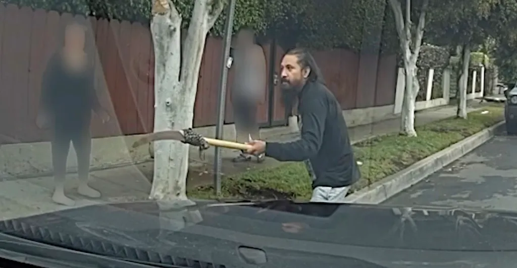 Bodycam footage shows a man holding a pole saw and approaching a police vehicle on a street.