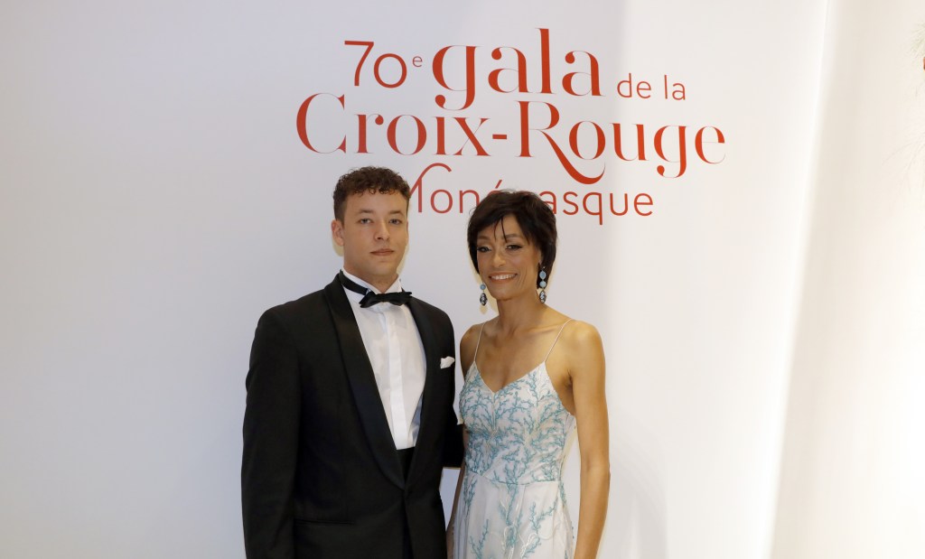 Antoine de Massy posing in a tuxedo alongside a woman in a light-colored dress in front of a red-lettered backdrop.