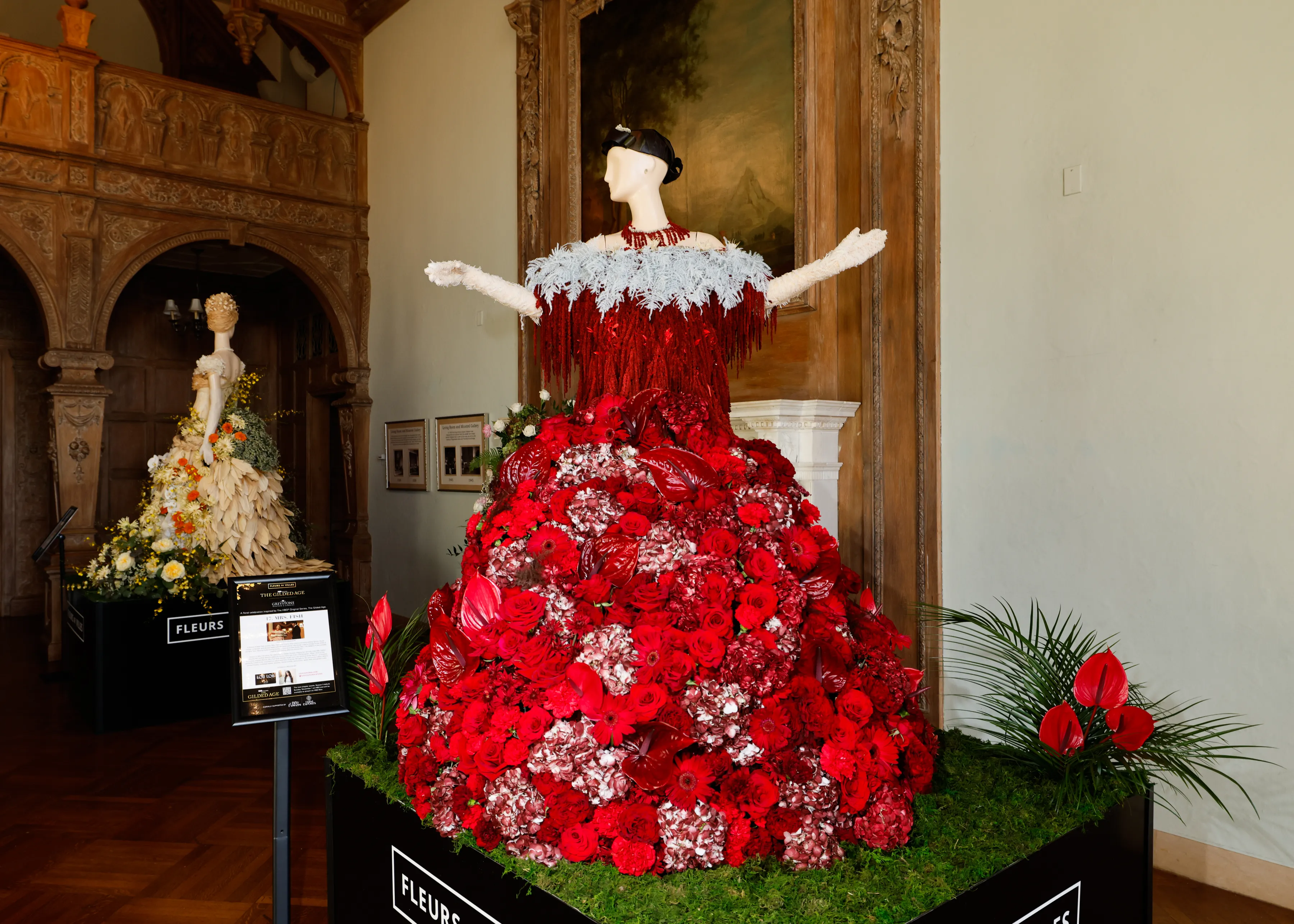A mannequin wearing a gown made of red and white flowers stands in a grand room with another floral sculpture in the background.