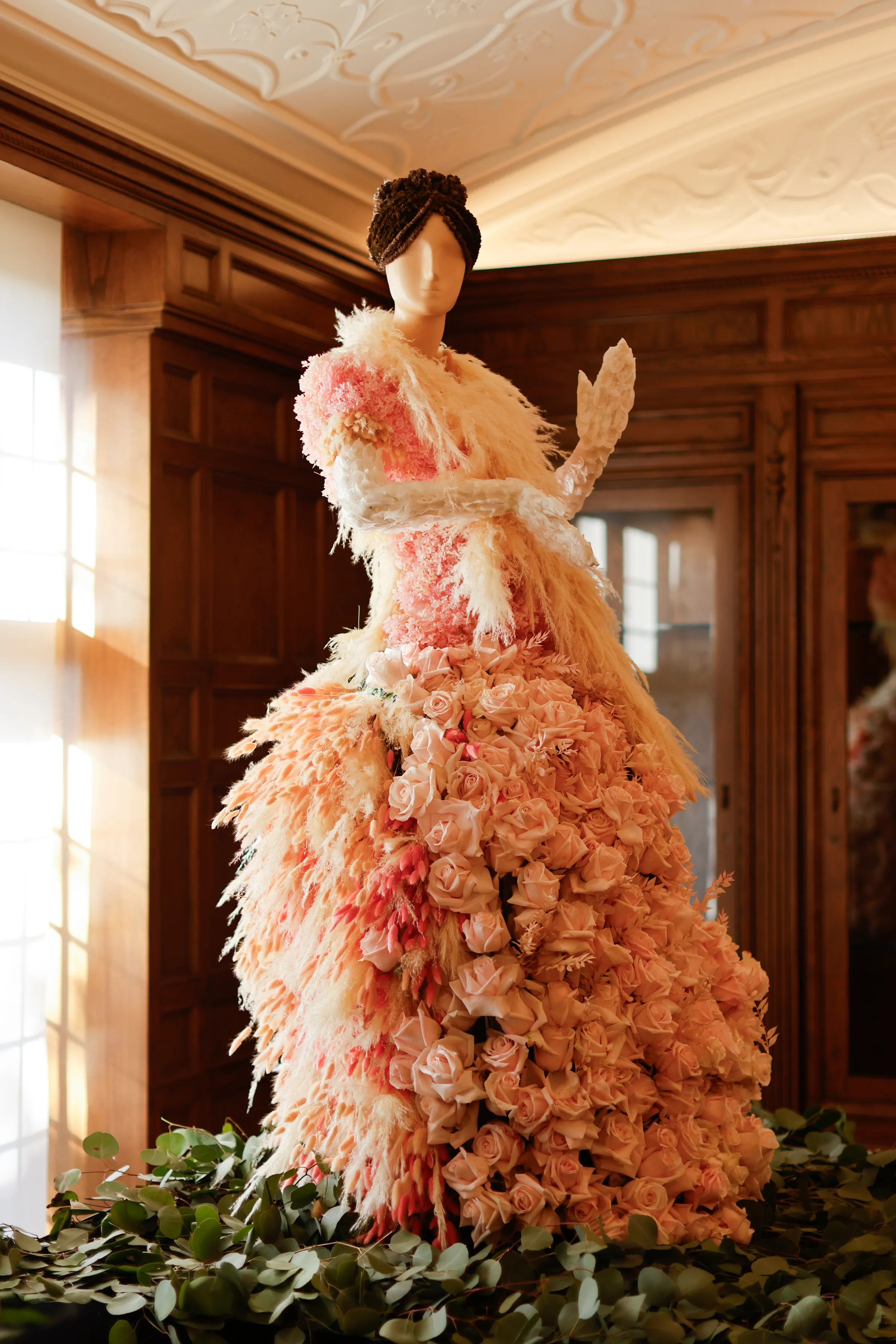 A mannequin wearing a floral gown made of pink roses and pampas grass stands indoors.