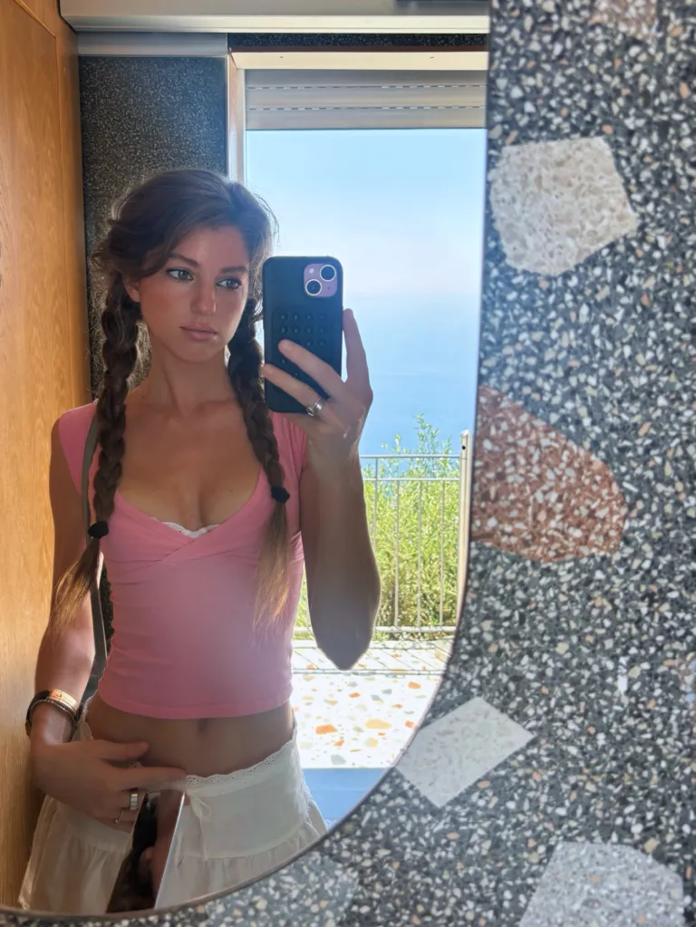 A young woman taking a mirror selfie, wearing a pink top and white skirt with her hair in two braids.