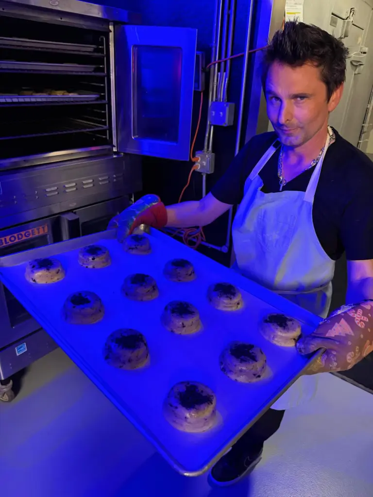 Muse frontman Matt Bellamy wearing an apron and oven mitts, holding a baking sheet of cookies in front of an oven.