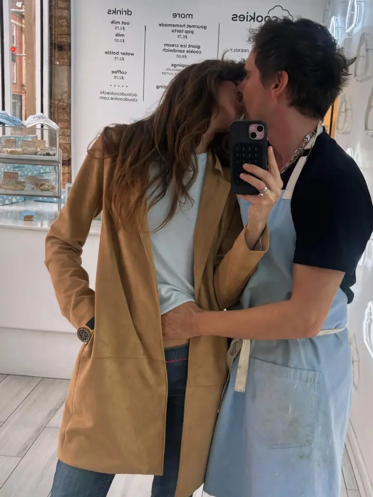 Matt Bellamy and a woman kissing while taking a mirror selfie inside a bakery.