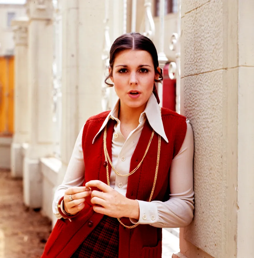 Portrait of Susan Saint James leaning against a stone pillar, wearing a red sweater vest and a white button-down shirt.