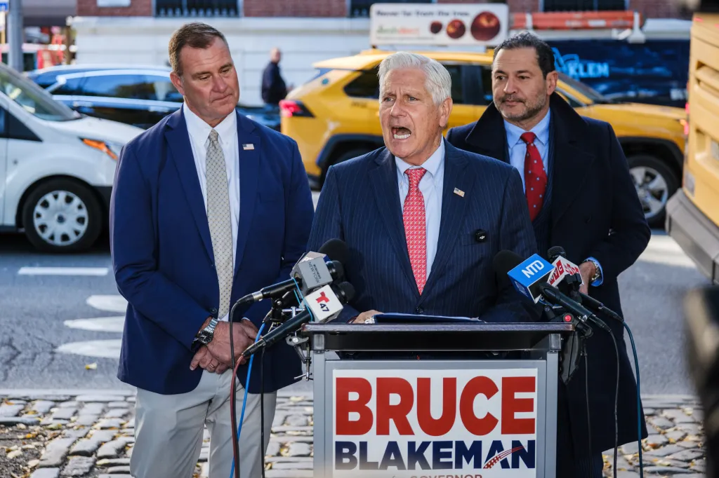 Bruce Blakeman speaks at a campaign event, flanked by supporters and microphones.