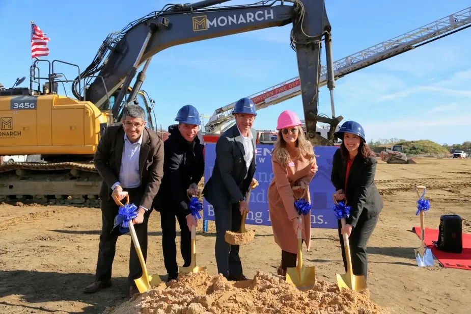 Five people in hard hats hold gold shovels with blue ribbons for a groundbreaking ceremony at an abandoned lot, with a large excavator in the background.