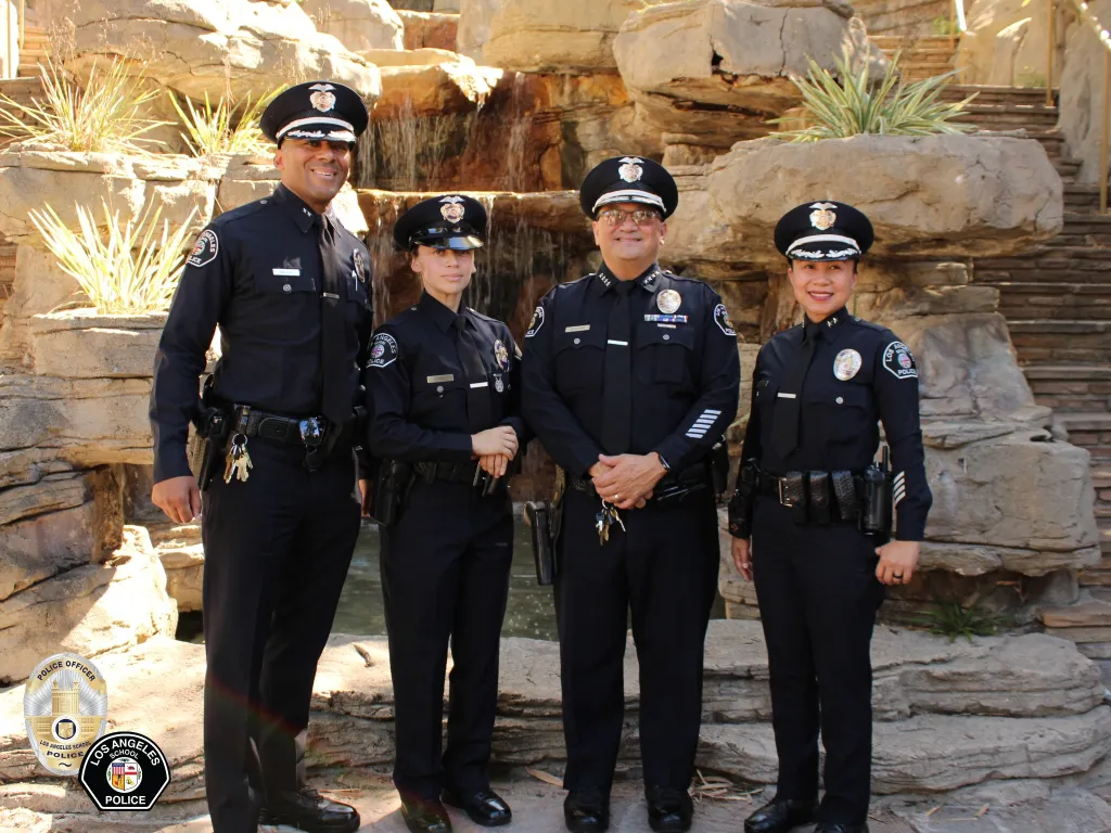 Four officers of the Los Angeles School Police Department in uniform pose in front of a rock and water feature.