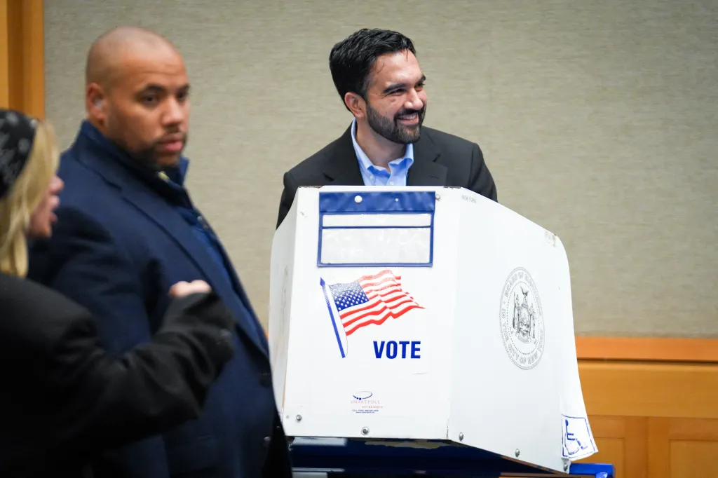 New York City Mayor Zohran Mamdani smiles behind a voting booth decorated with an American flag and the word 