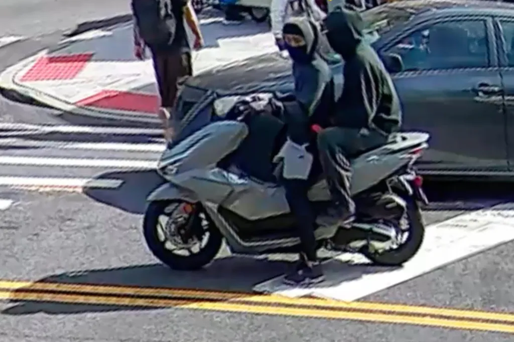 Two individuals wearing dark hooded sweatshirts and face coverings ride a light-colored scooter on a city street near a car.