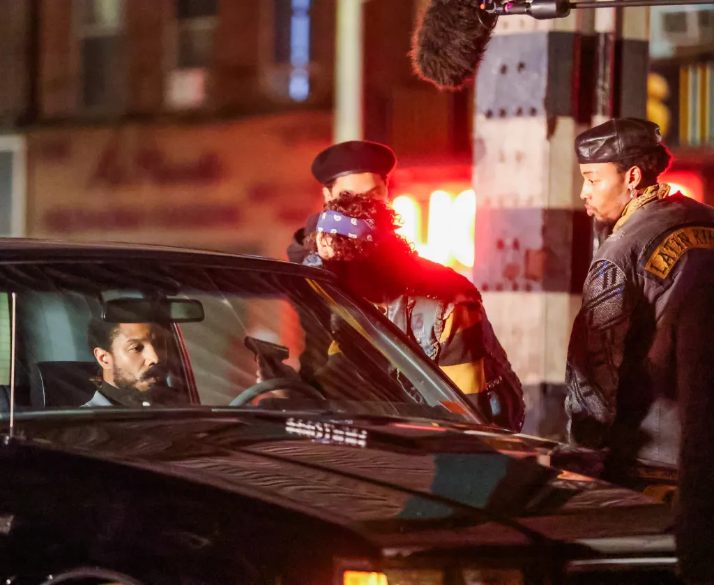 Michael B. Jordan sits inside a car at night while a person wearing a blue bandana holds a gun near his face during the filming of 