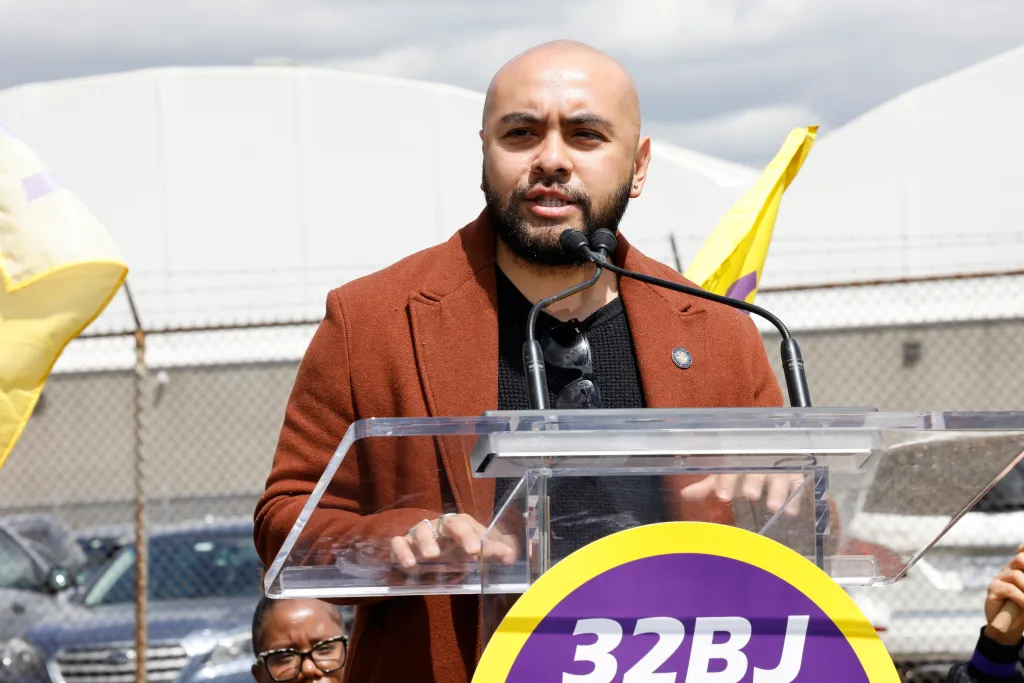 New York State Assemblymember Steven Raga speaks into microphones at a podium during a protest at JFK Airport.
