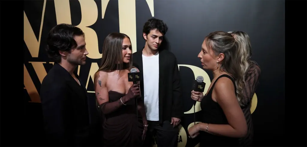Ariana Biermann and Liam Obergfoll being interviewed by reporters in front of a black backdrop with gold lettering.