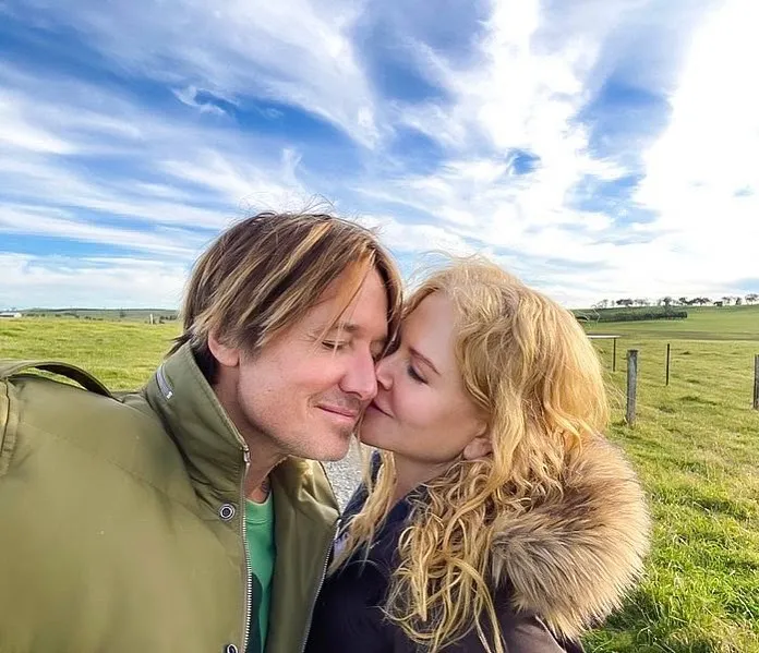 Nicole Kidman, in a jacket with fur collar, kisses Keith Urban, in a green jacket, on the cheek in a grassy field under a cloudy blue sky.