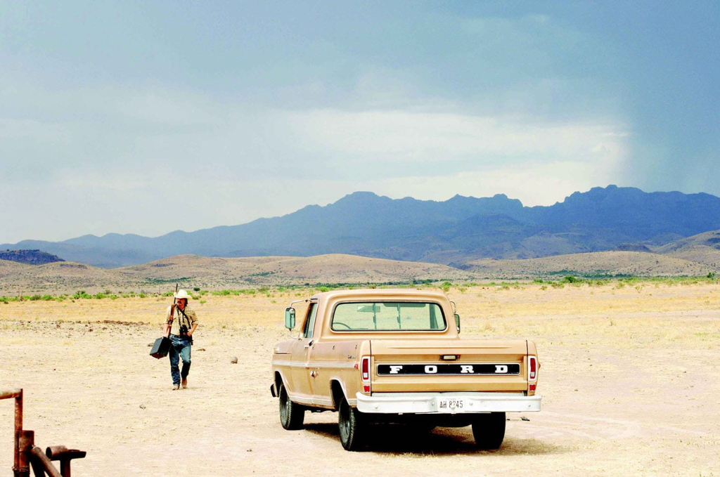 A man walking across a desert landscape in front of a parked truck with mountains in the background.
