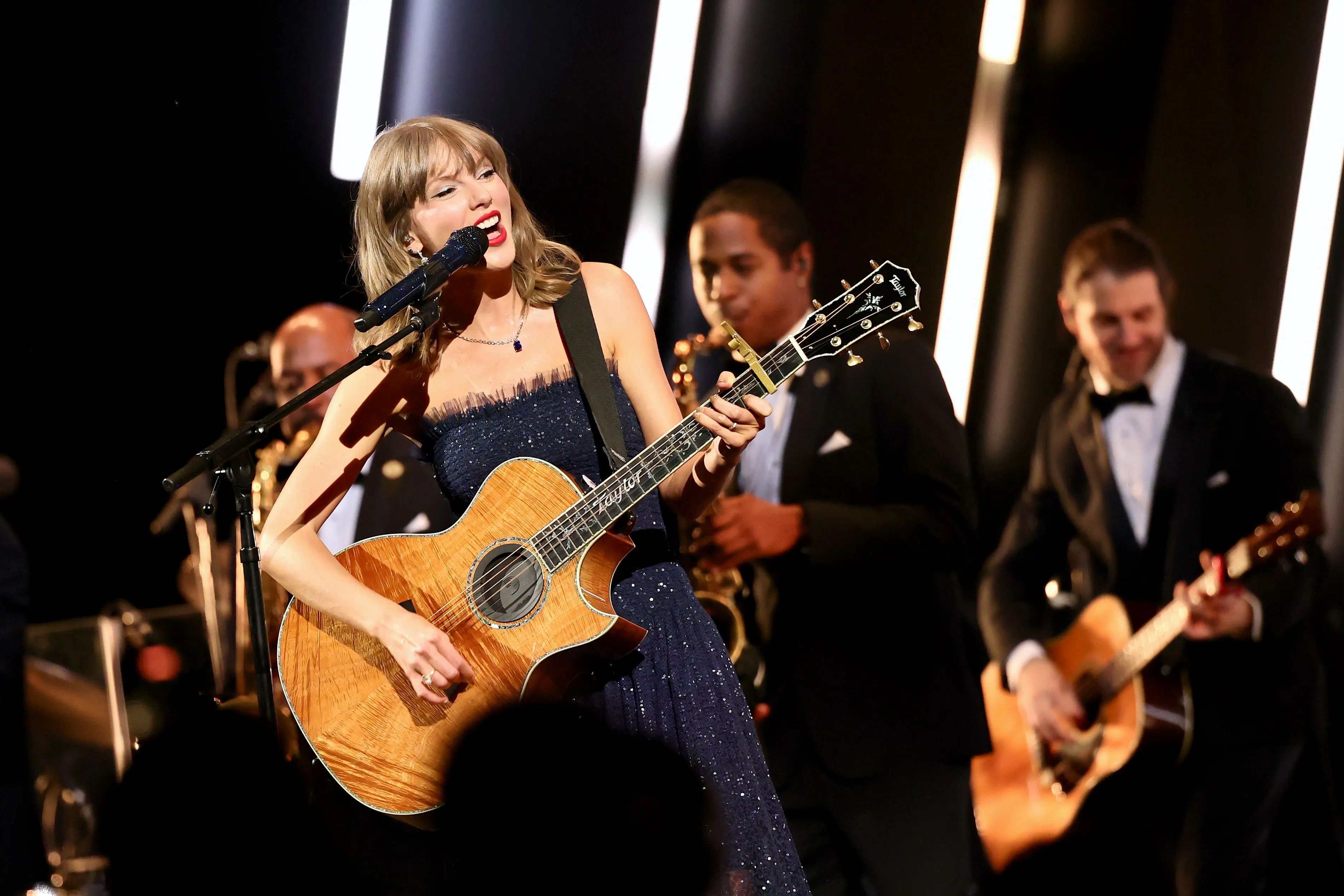 Taylor Swift performs onstage with an acoustic guitar, accompanied by a band.