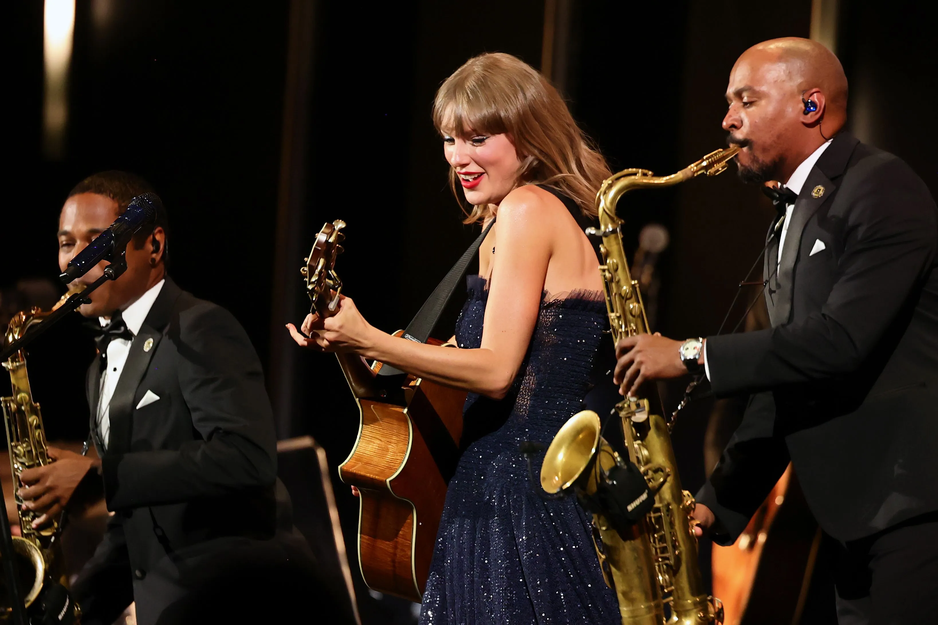 Taylor Swift performs onstage, playing acoustic guitar flanked by band members playing saxophones during the Academy Museum Gala.