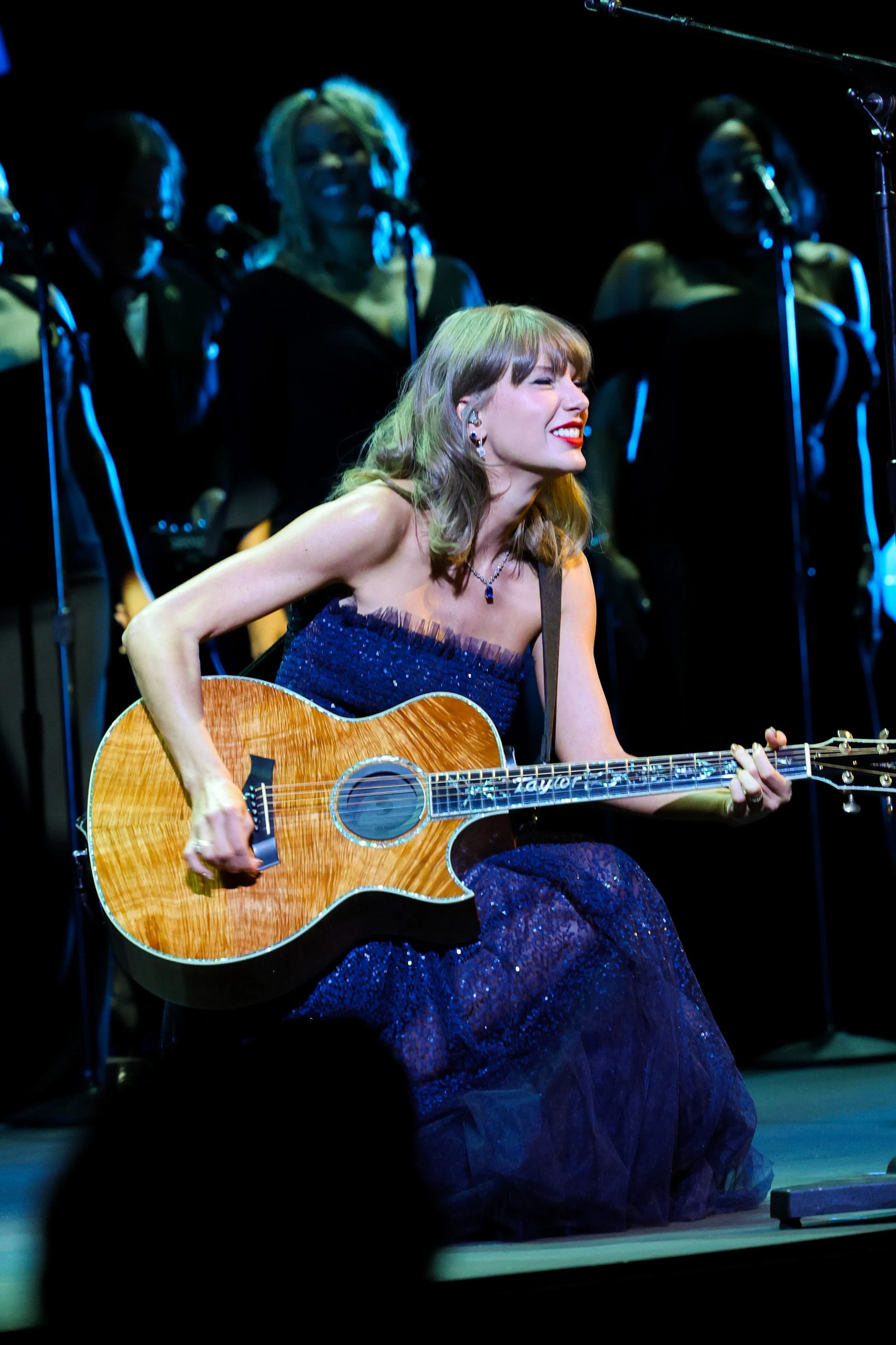 Taylor Swift plays an acoustic guitar while performing on stage in a dark blue gown.