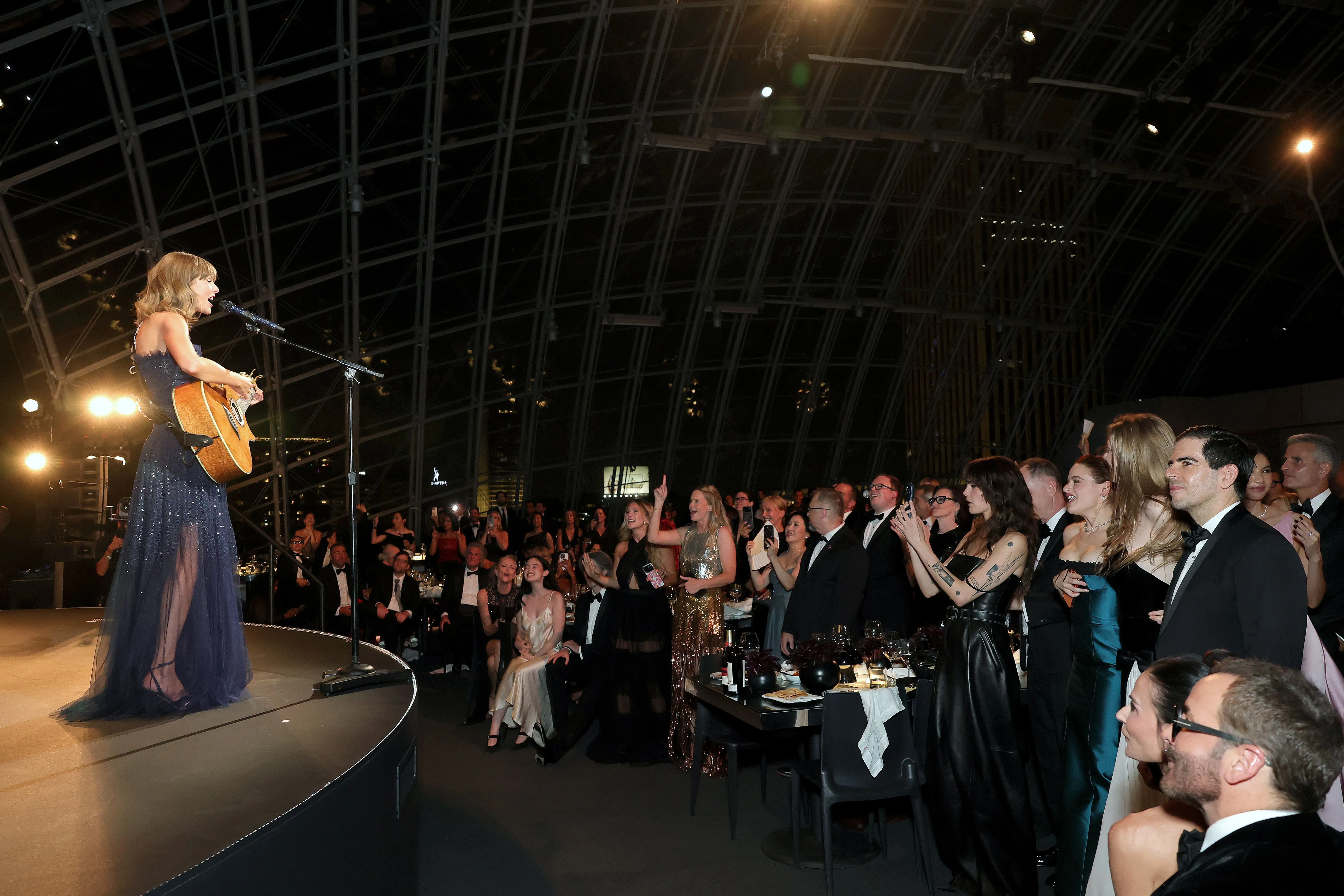 Taylor Swift performs on stage with a guitar at the Academy Museum Gala, as an audience seated at tables looks on.