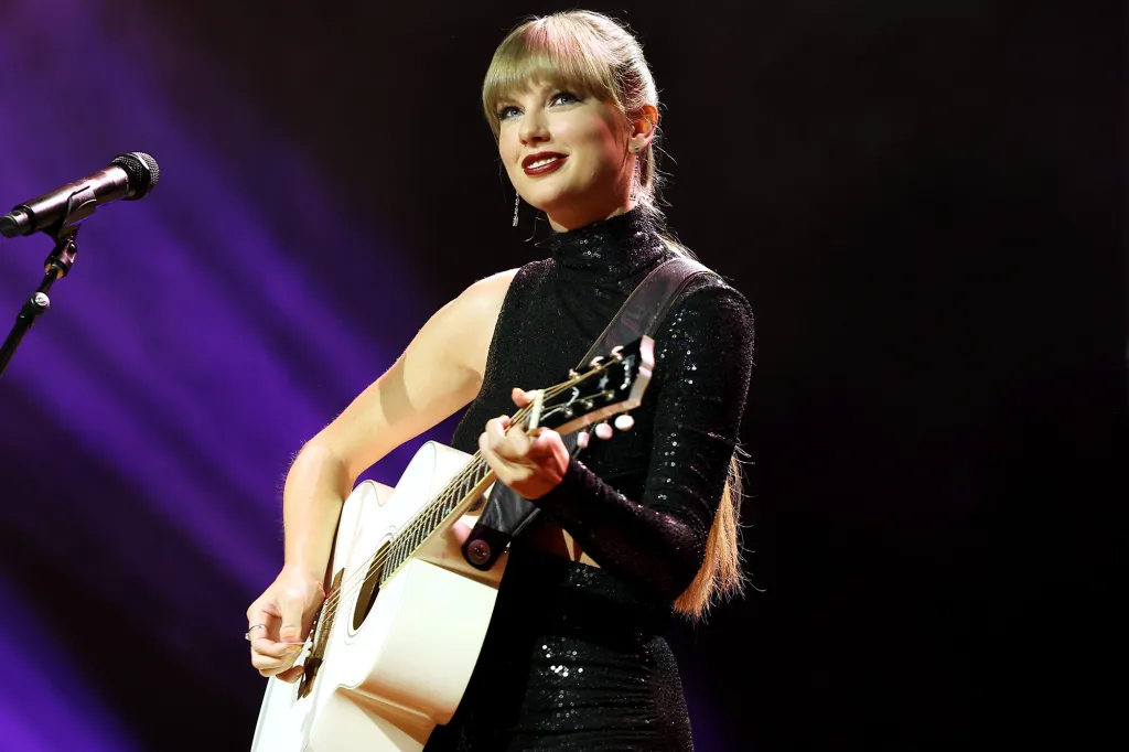Taylor Swift performs onstage, playing an acoustic guitar and singing into a microphone.
