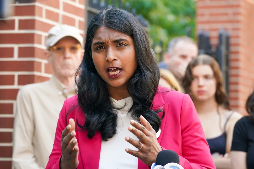 New York Attorney General candidate Saritha Komatireddy speaks outdoors with her hands raised in front of a brick wall, with several residents in the background.