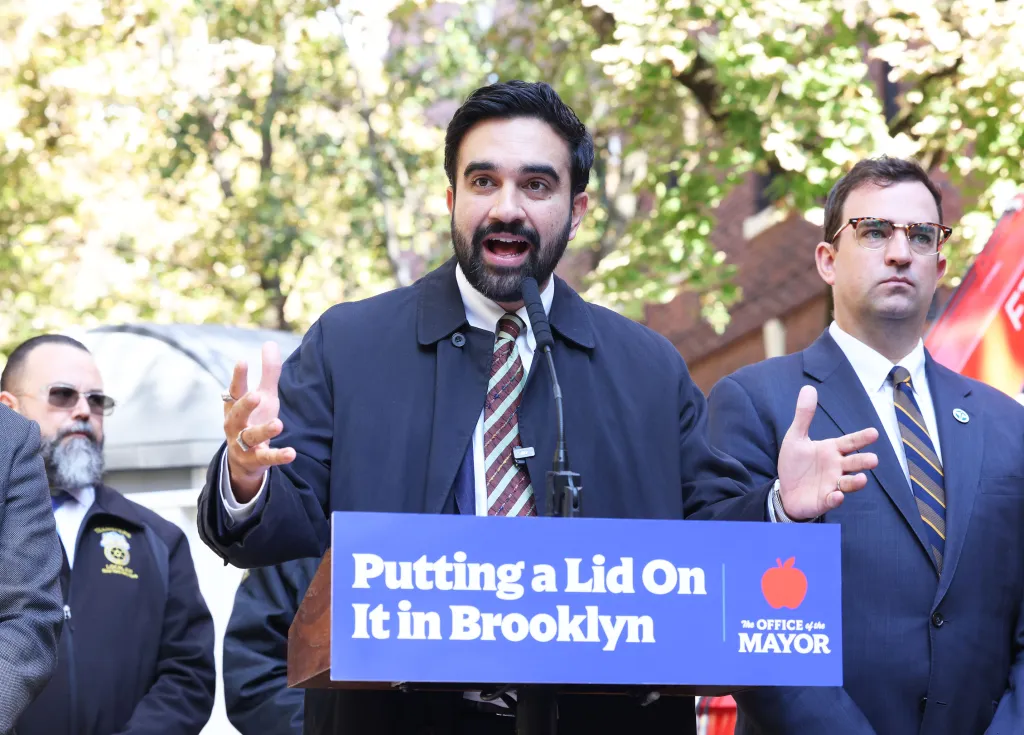 NYC Mayor Zohran Mamdani speaks at a podium with Sanitation Commissioner Gregory Anderson standing beside him in Brooklyn, NY.