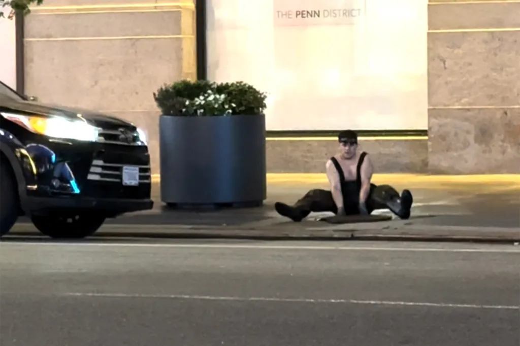 A person sitting next to an open manhole on a New York City sidewalk.