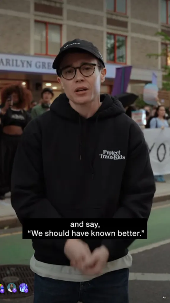 Elliot Page stands outside a building in front of a crowd, wearing a black hoodie that reads 