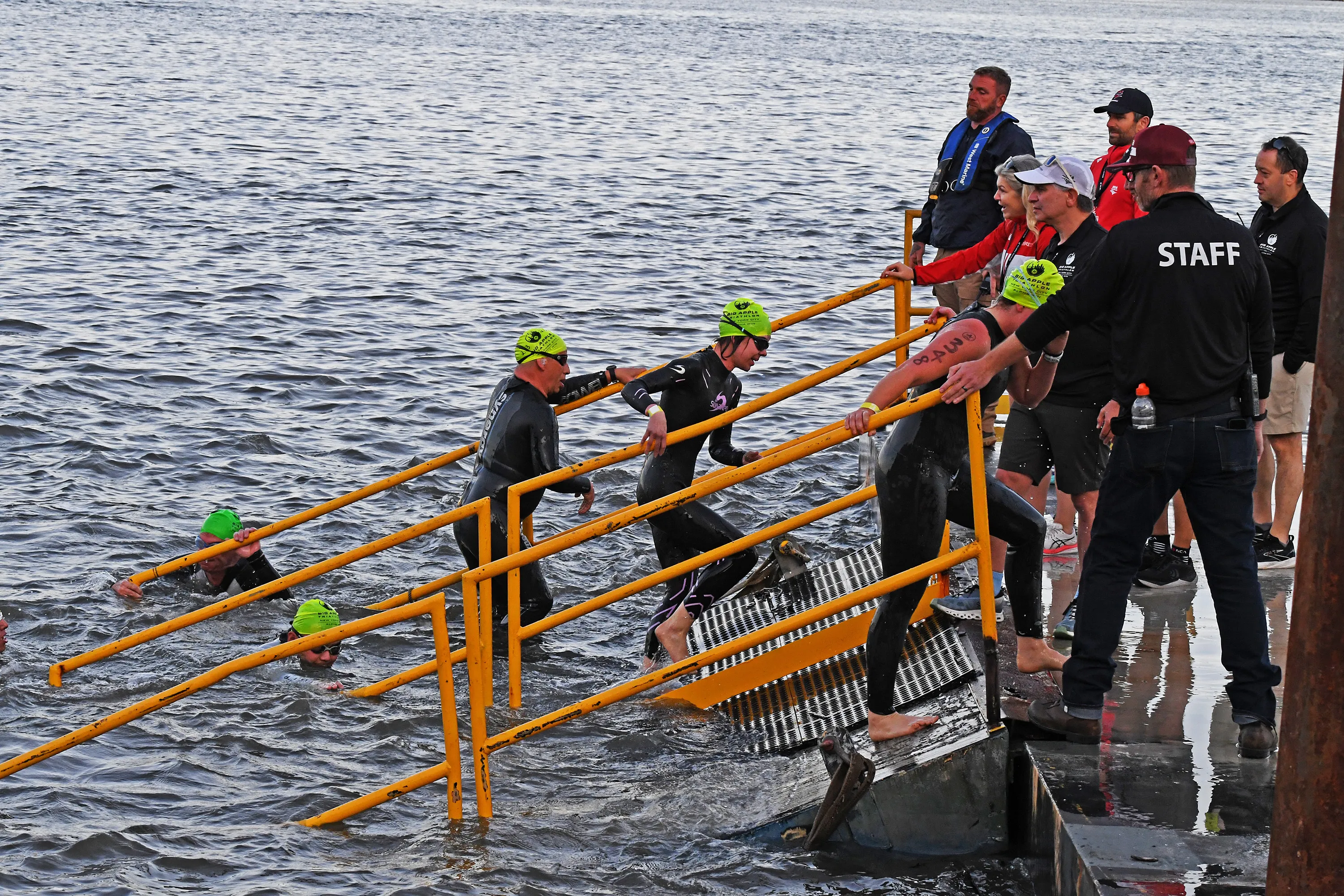Triathletes exit the water using a yellow railing and metal steps during the NYC Triathlon.