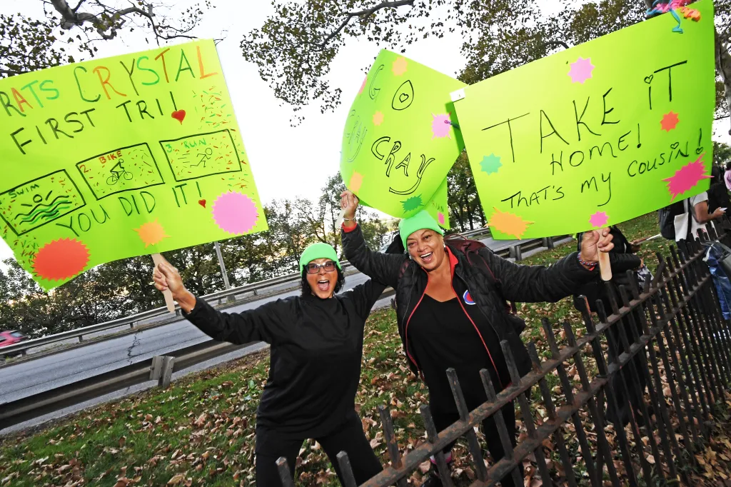 Two smiling women hold up bright green signs cheering on competitors at the NYC Triathlon.