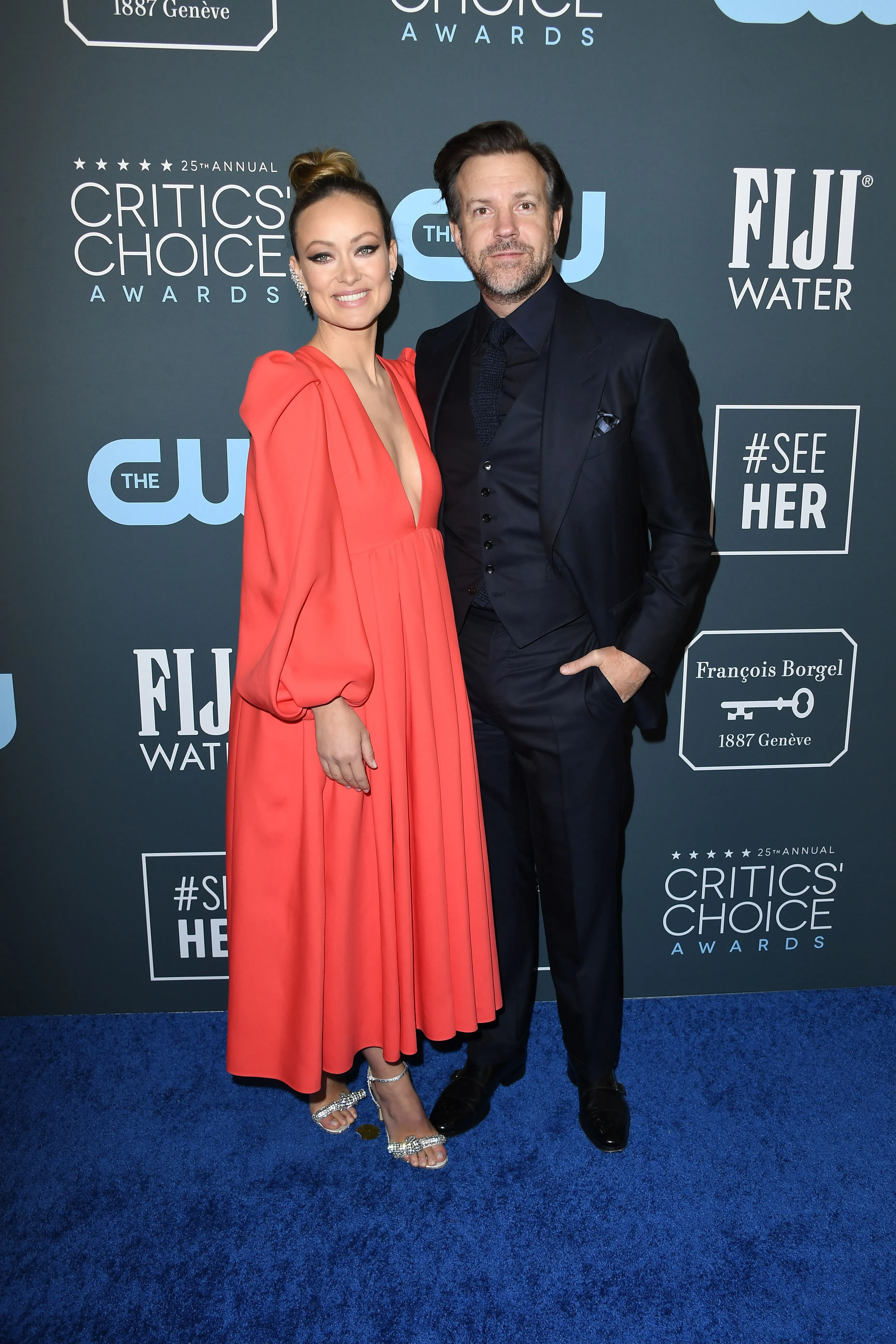 Olivia Wilde in a long red dress and Jason Sudeikis in a dark suit pose on the blue carpet at the Critics' Choice Awards.