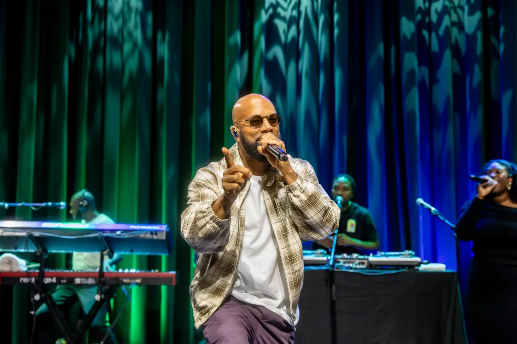 Common performing on stage with a backing band and singers in front of green and blue curtains.