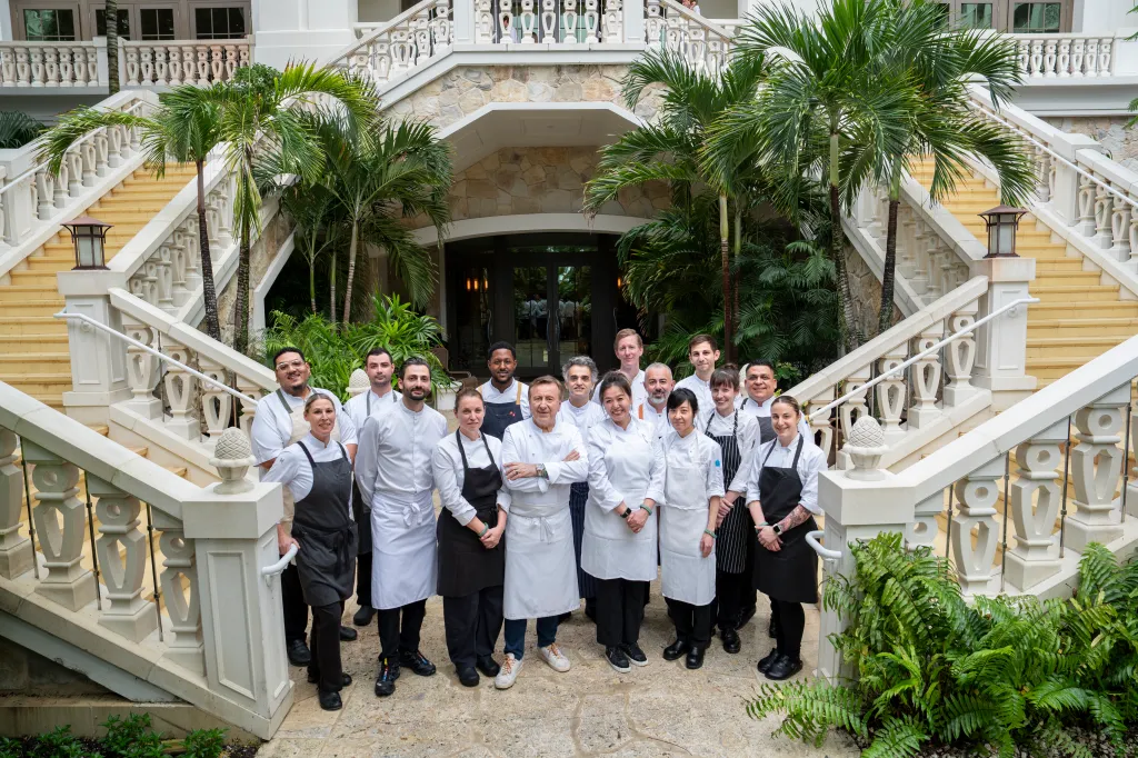 Chef Daniel Boulud and alumni chefs pose outside in front of a grand white staircase and palm trees.