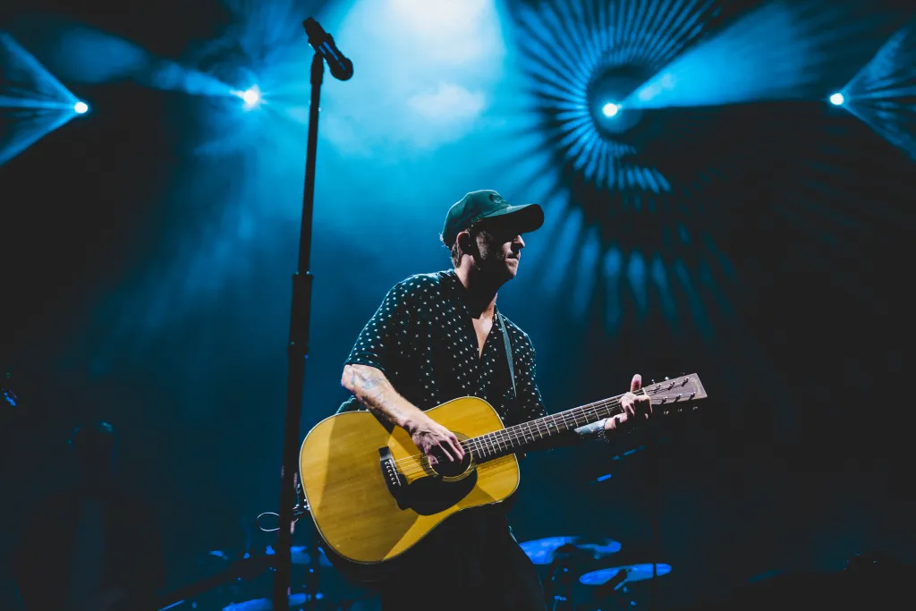 Ryan Tedder of OneRepublic playing an acoustic guitar on stage under blue stage lighting.