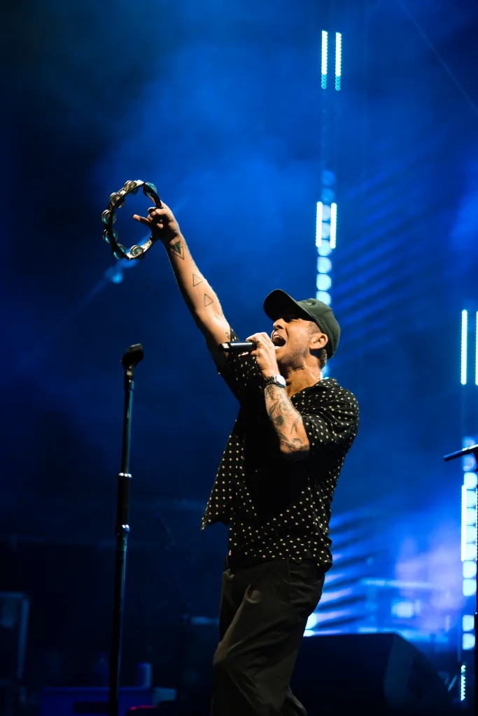 Ryan Tedder of OneRepublic performing live on stage, holding a microphone and a tambourine under blue stage lighting.