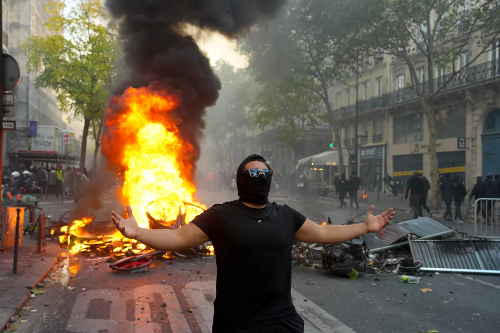 A protester with face covered and arms outstretched stands in front of a burning barricade.