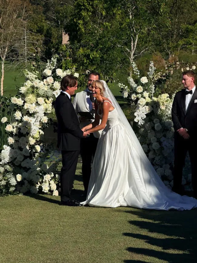 Annie Knight and Henry Brayshaw smiling at their wedding ceremony, surrounded by white floral arrangements.
