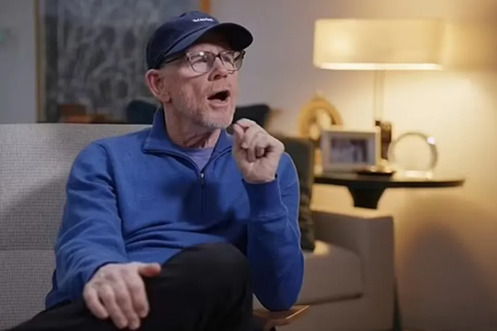 Ron Howard speaking during an interview in a blue sweater, baseball cap, and glasses.
