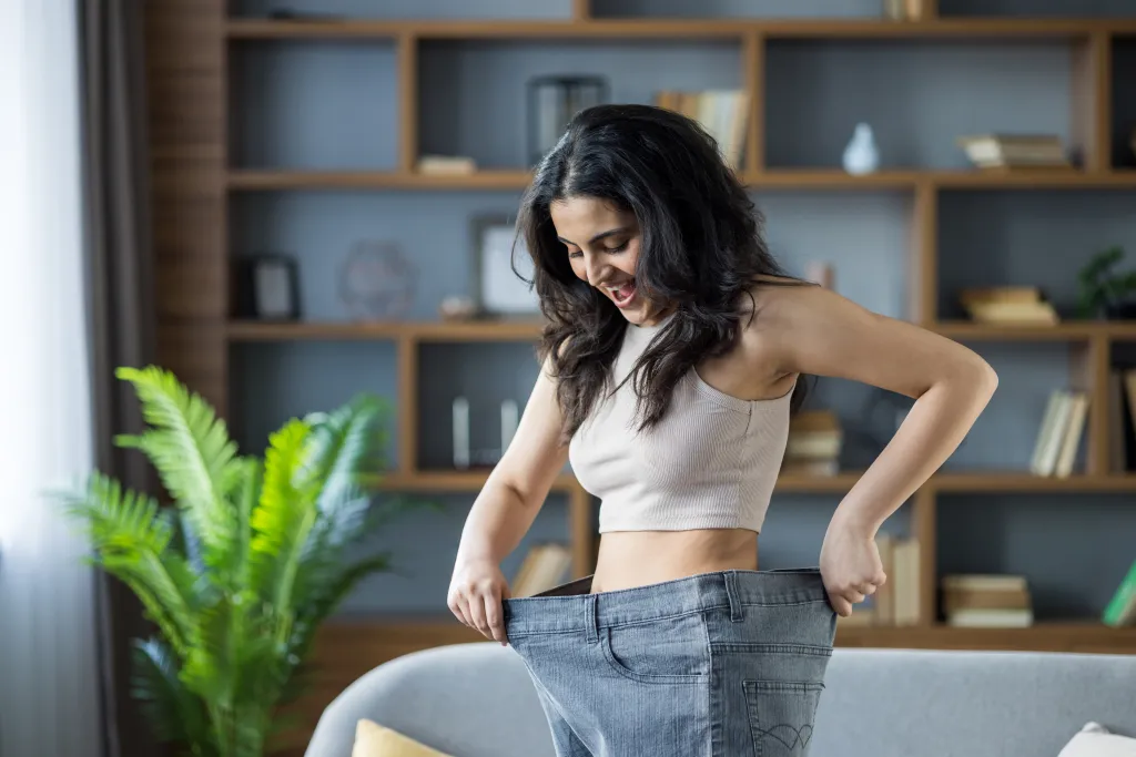 A young woman smiling and holding out her oversized jeans to show her successful weight loss.