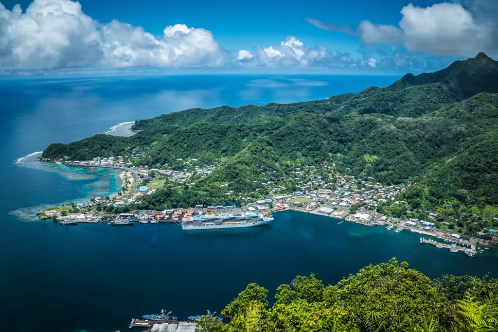 An elevated view of Pago Pago, American Samoa, shows a large cruise ship docked in the bay, surrounded by lush green hills and the blue Pacific Ocean.