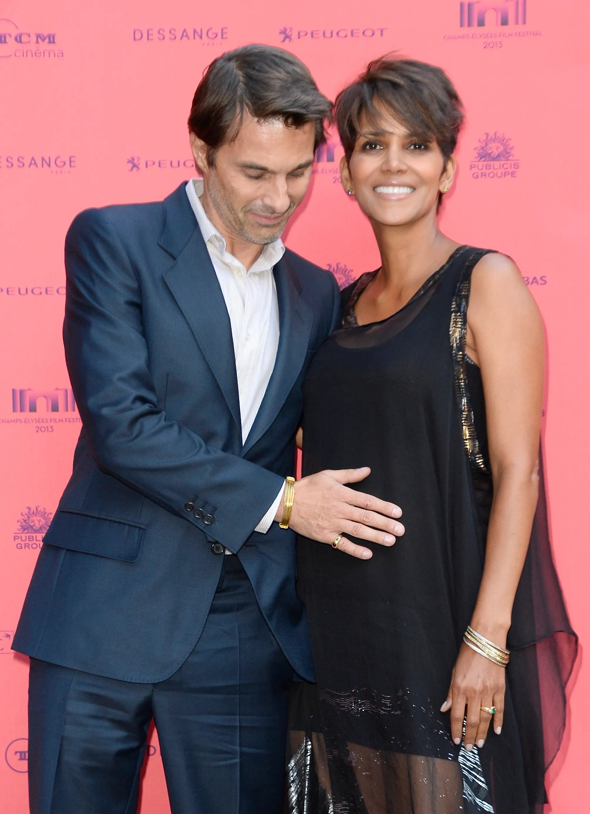 Halle Berry and Olivier Martinez stand together on a red carpet, with Martinez smiling down and placing his hand on Berry's pregnant abdomen.
