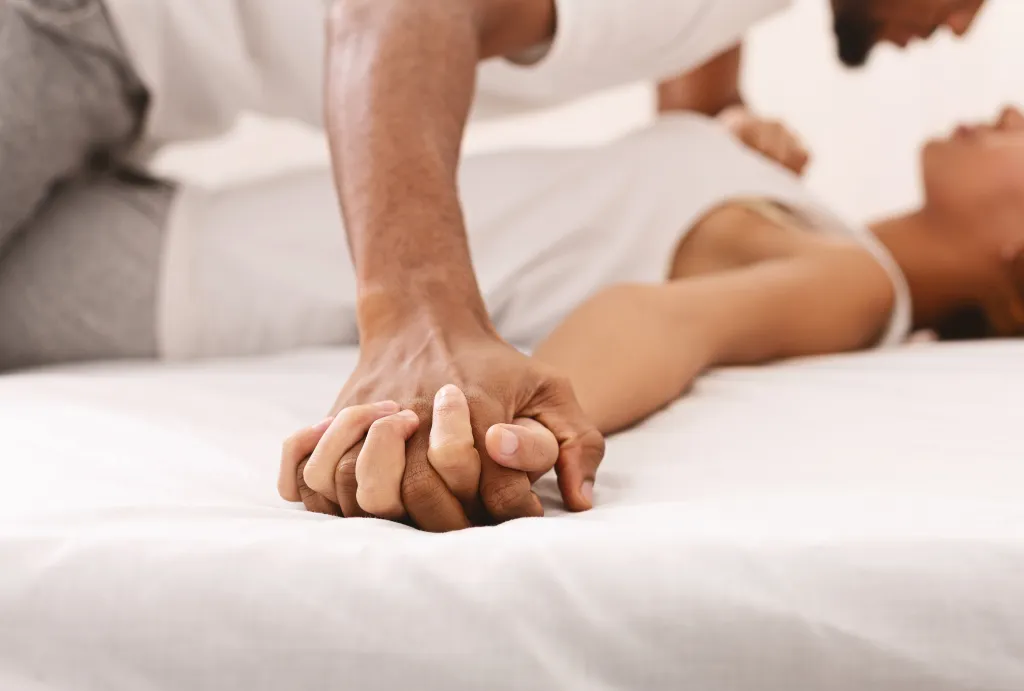An African-American couple holding hands in bed, focusing on their intertwined hands in the foreground.
