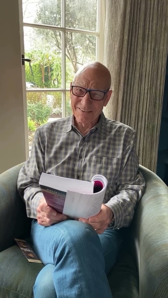 Patrick Stewart sitting in a chair holding a book and smiling.